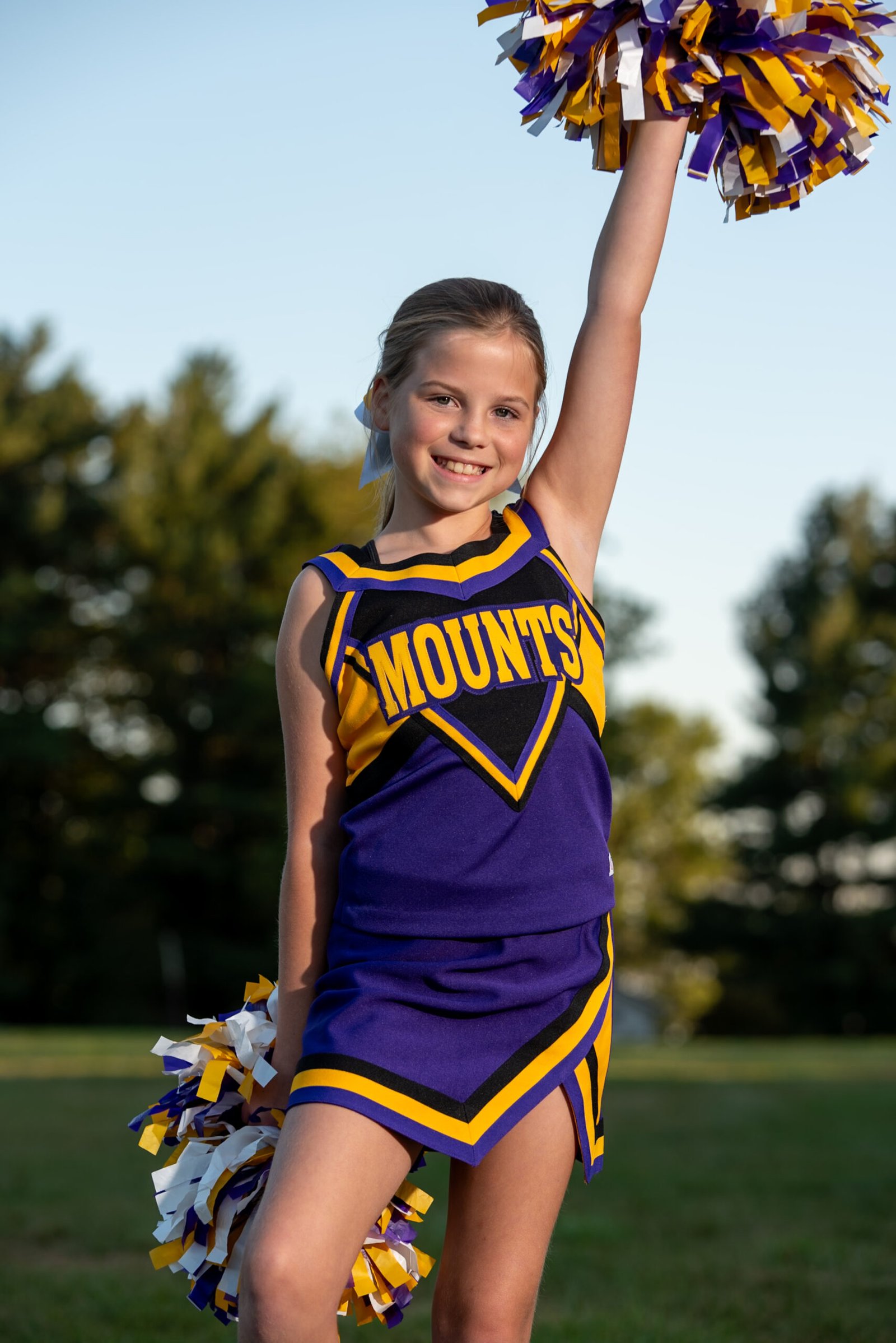 Youth cheerleader in a Mounts uniform posing outdoors with one arm raised during golden hour in Ephrata, PA.