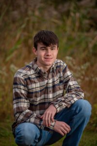High school senior sitting in a grassy fall field wearing a plaid flannel shirt and jeans during an outdoor portrait session in Ephrata, PA
