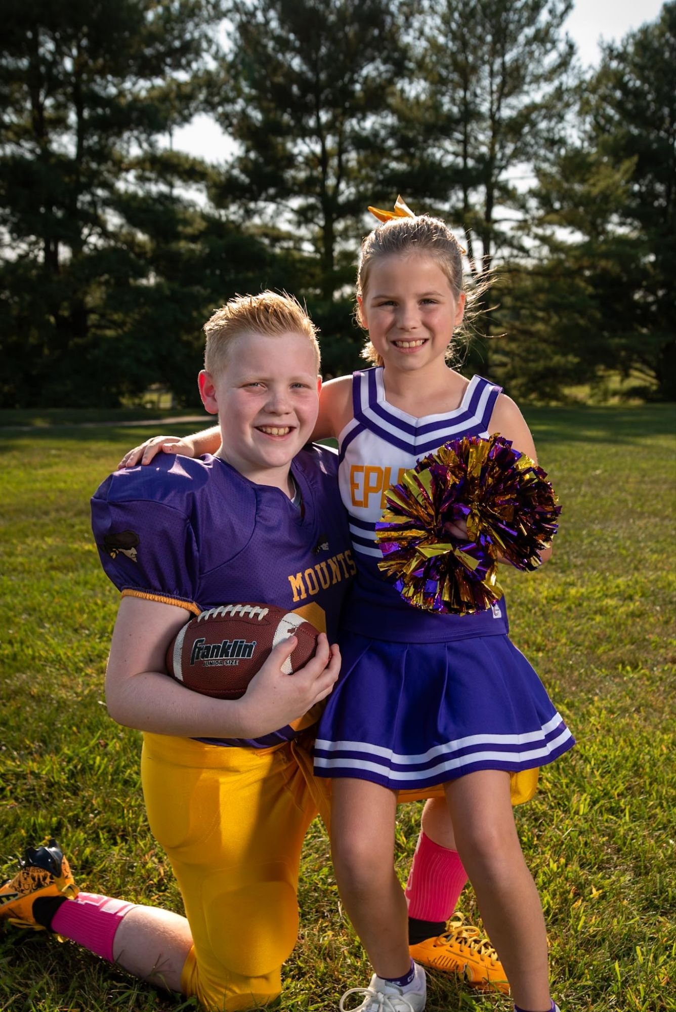 A young football player and a cheerleader standing together on a grassy field, the cheerleader holding pom-poms