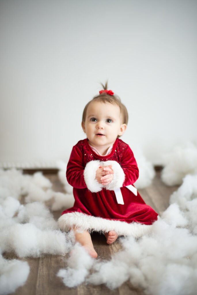 Baby girl in a red and white Christmas dress sitting on a studio floor, smiling with soft natural light in Ephrata, PA