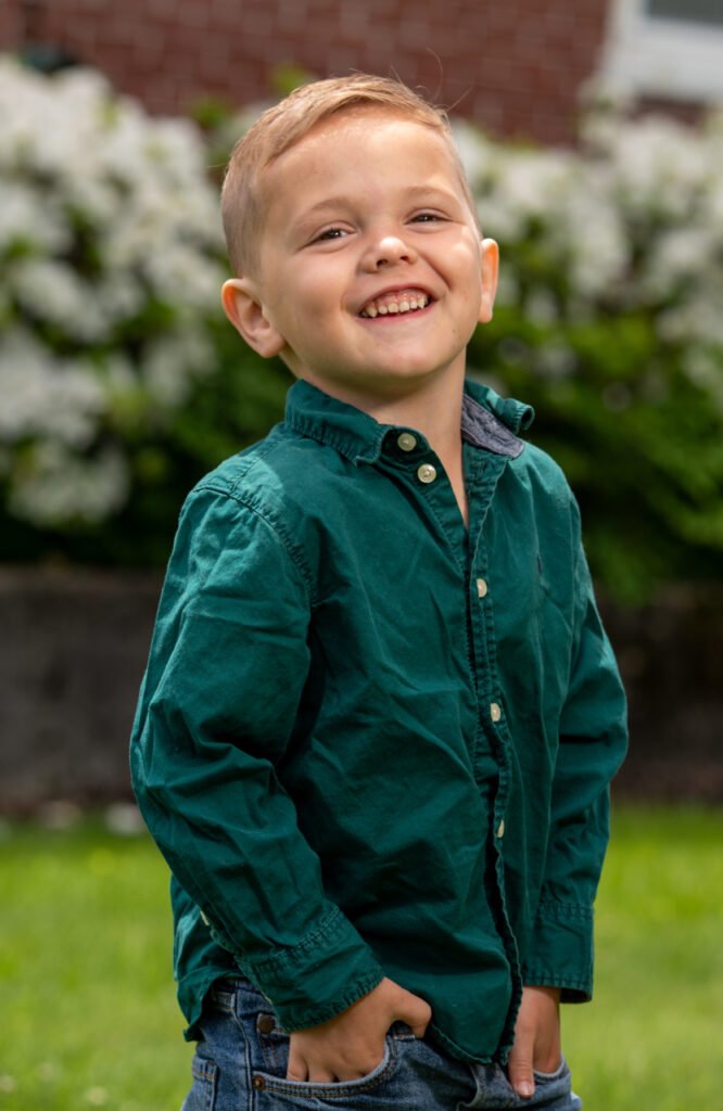 Candid outdoor portrait of a little boy in a green shirt smiling with greenery in the background in Ephrata, PA.