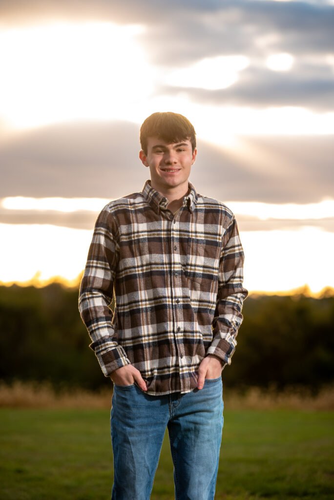 High school senior standing in a grassy field during golden hour with sun rays and clouds behind him, outdoor portrait in Ephrata, PA.