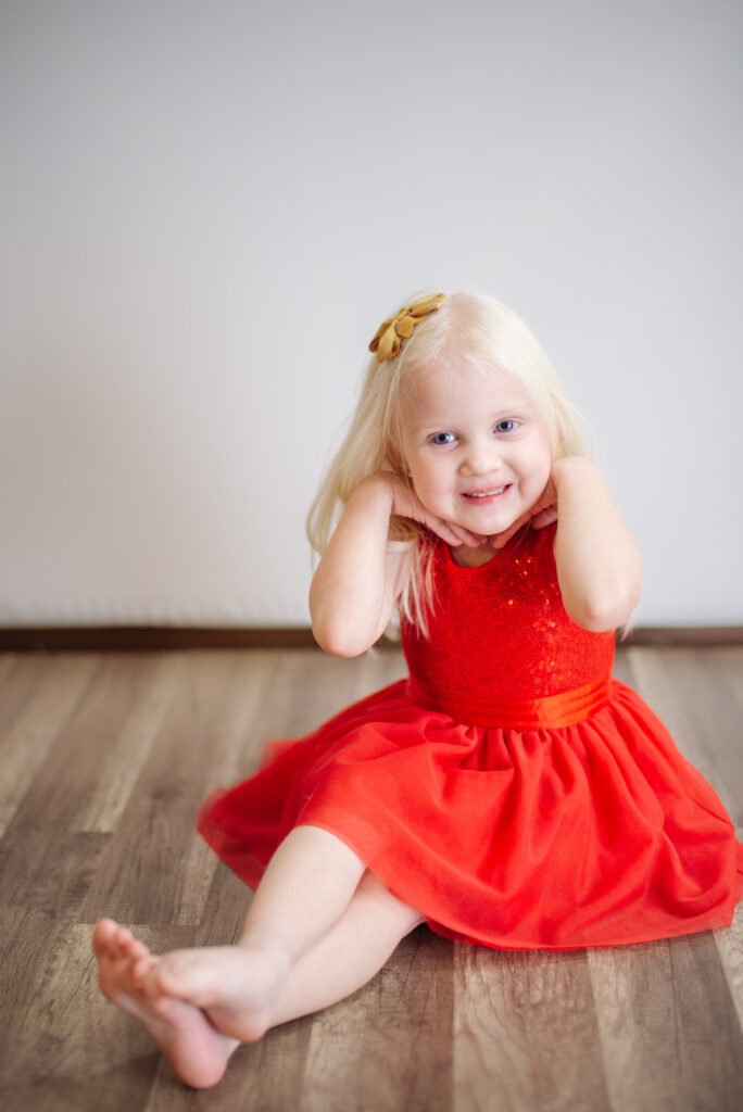 Toddler girl in a bright red dress sitting on a studio floor, smiling with soft natural light in Ephrata, PA.