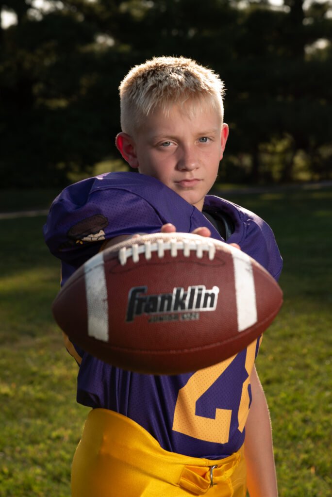 Youth football player in a purple jersey holding a football toward the camera during an outdoor portrait in Ephrata, PA.